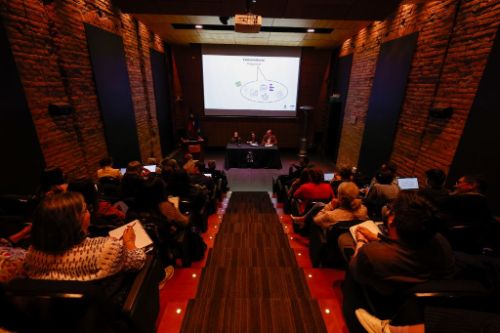Auditorio Facultad de Arquitectura y Urbanismo desde atrás. Se ve a las personas sentadas y adelante una mesa en que conversan las profesoras Rubliar y Armanet, además del profesor Olea.